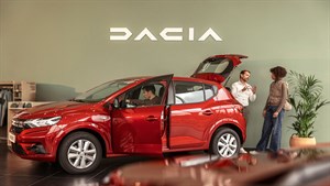 Crvena Dacia u salonu s otvorenim vratima, dok troje ljudi razgovara pored vozila.








