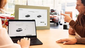 Dvoje ljudi sjedi za stolom u autosalonu i pregledava konfiguraciju vozila marke Dacia na prijenosnom računalu i većem ekranu ispred sebe.

