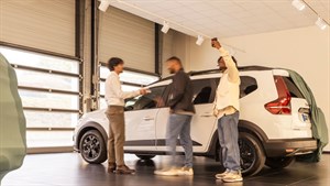 Trojica muškaraca stoje u salonu pokraj bijelog SUV vozila marke Dacia Jogger. Jedan od njih se rukuje s prodavačem, dok drugi fotografira vozilo mobitelom.

