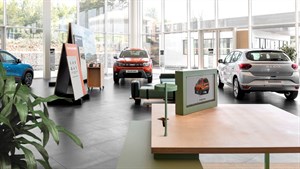 Unutrašnjost moderne prodajne kuće Dacia s velikim staklenim stijenama, prikazuje izložena vozila uključujući Dacia Bigster, Sandero i Duster, uz informacijske panele i kutak za korisnike.


