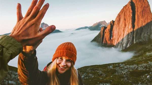 Žena s narančastom kapom nasmijano podiže ruku prema osobi ispred sebe, s planinama i morem oblaka u pozadini.