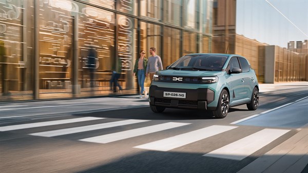 Dacia Spring u plavoj boji vozi gradskom ulicom, snimljen sprijeda, dok pješaci prolaze uz modernu urbanu zgradu.






