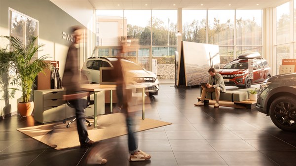 Prikaz modernog Dacia salona s nekoliko vozila, kupcima u pokretu i osobom koja sjedi u kutku salona.