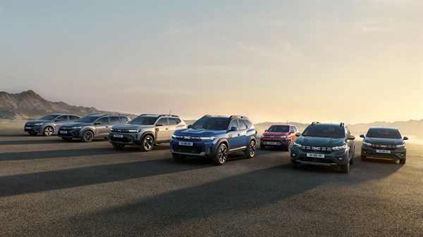 Više Dacia vozila parkiranih jedno uz drugo na otvorenom prostoru s planinskom pozadinom pri zalasku sunca.

