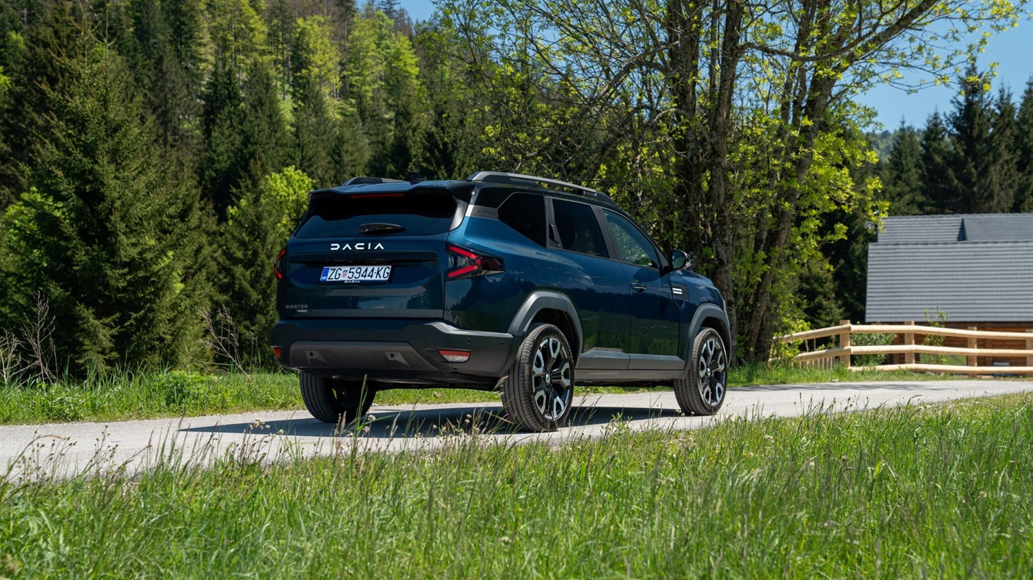 Tamnoplavi Dacia Bigster snimljen straga dok vozi seoskom cestom, okružen zelenilom, drvećem i kućom u pozadini.