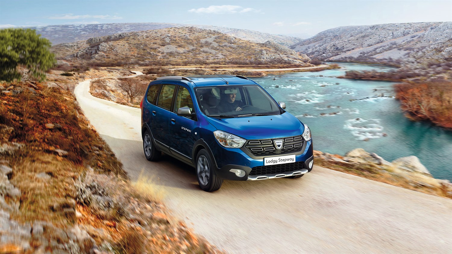 Plavi Dacia Lodgy u vožnji zavojitom cestom uz jezero i kameniti krajolik.