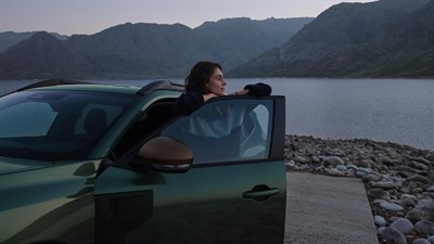 Žena naslonjena na otvorena vrata vozila Dacia u prirodi, uz jezero i planinski krajolik u pozadini.