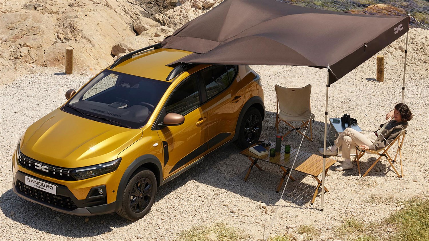 Žuto vozilo Dacia Sandero Stepway parkirano u prirodi, s osobom koja se odmara ispod tende uz opremu za kampiranje