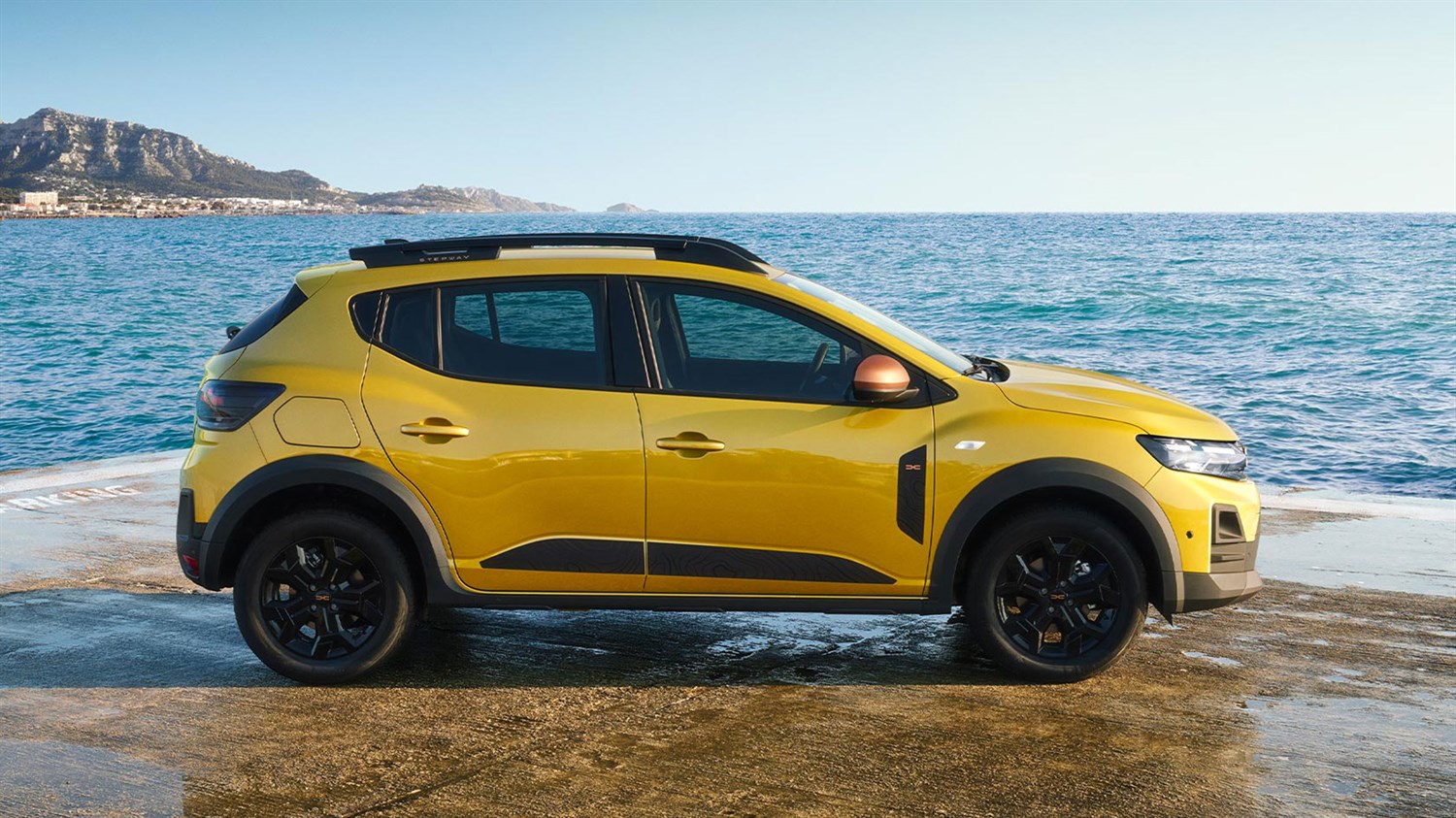 Žuto vozilo Dacia Sandero Stepway parkirano uz more, s planinskim krajolikom u pozadini.