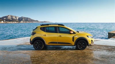 Bočni pogled na žuto Dacia vozilo parkirano uz more, s planinama u pozadini.