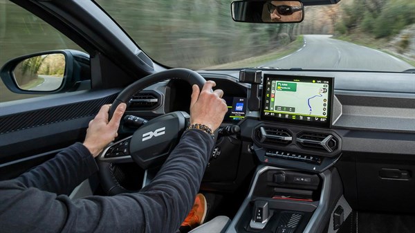 Vozač upravlja plavim vozilom Dacia po zavojitoj cesti kroz šumoviti krajolik, uz navigaciju prikazanu na središnjem zaslonu.