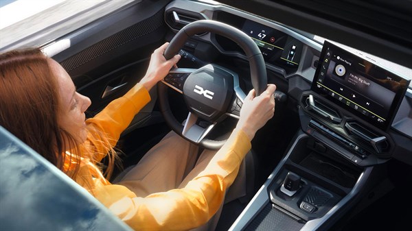 Žena u narančastoj košulji upravlja vozilom Dacia, s pogledom na digitalnu instrumentnu ploču i središnji multimedijski zaslon.