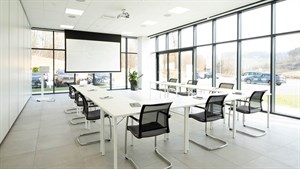 Svijetla i moderna konferencijska dvorana s velikim staklenim stijenkama, opremljena bijelim stolovima, crnim stolicama te projektorom s platnom.

