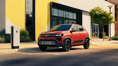 Crvena Dacia Spring Electric parkirana pored javne punionice, u urbanom okruženju ispred zgrade sa žuto-bijelom fasadom.