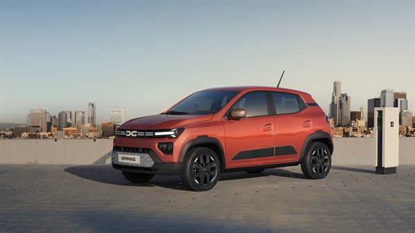 Crvena Dacia Spring Electric parkirana na otvorenom parkiralištu pored punionice, s panoramom grada u pozadini.