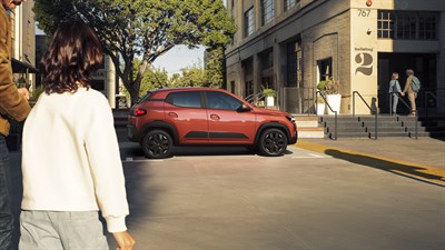 Crvena Dacia Spring Electric parkirana uz rub pločnika ispred zgrade, dok prolaznici šetaju u prvom planu.