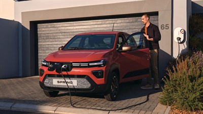 Crvena Dacia Spring Electric priključena na kućnu punionicu ispred garaže, dok vozač stoji pored otvorenih vozačevih vrata.