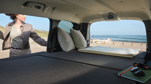 Unutrašnjost vozila Dacia Jogger s postavljenim krevetom i otvorenim stražnjim poklopcem uz pogled na plažu.