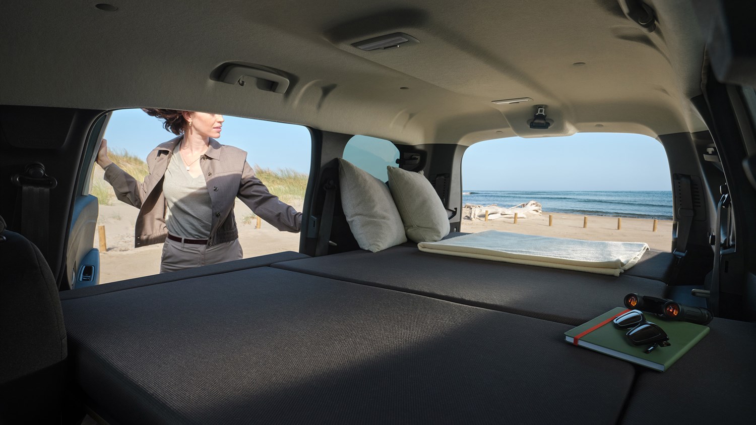 Unutrašnjost vozila Dacia Jogger s potpuno spuštenim stražnjim sjedalima koja stvaraju ravnu površinu nalik ležaju, s jastucima i bilježnicom na stražnjem dijelu, uz pogled na plažu kroz otvorena stražnja vrata.