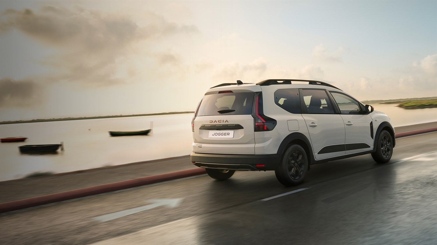 Bijelo vozilo Dacia Jogger prikazano sa stražnje strane u vožnji uz obalu, na cesti uz mirno more i brodice u pozadini.