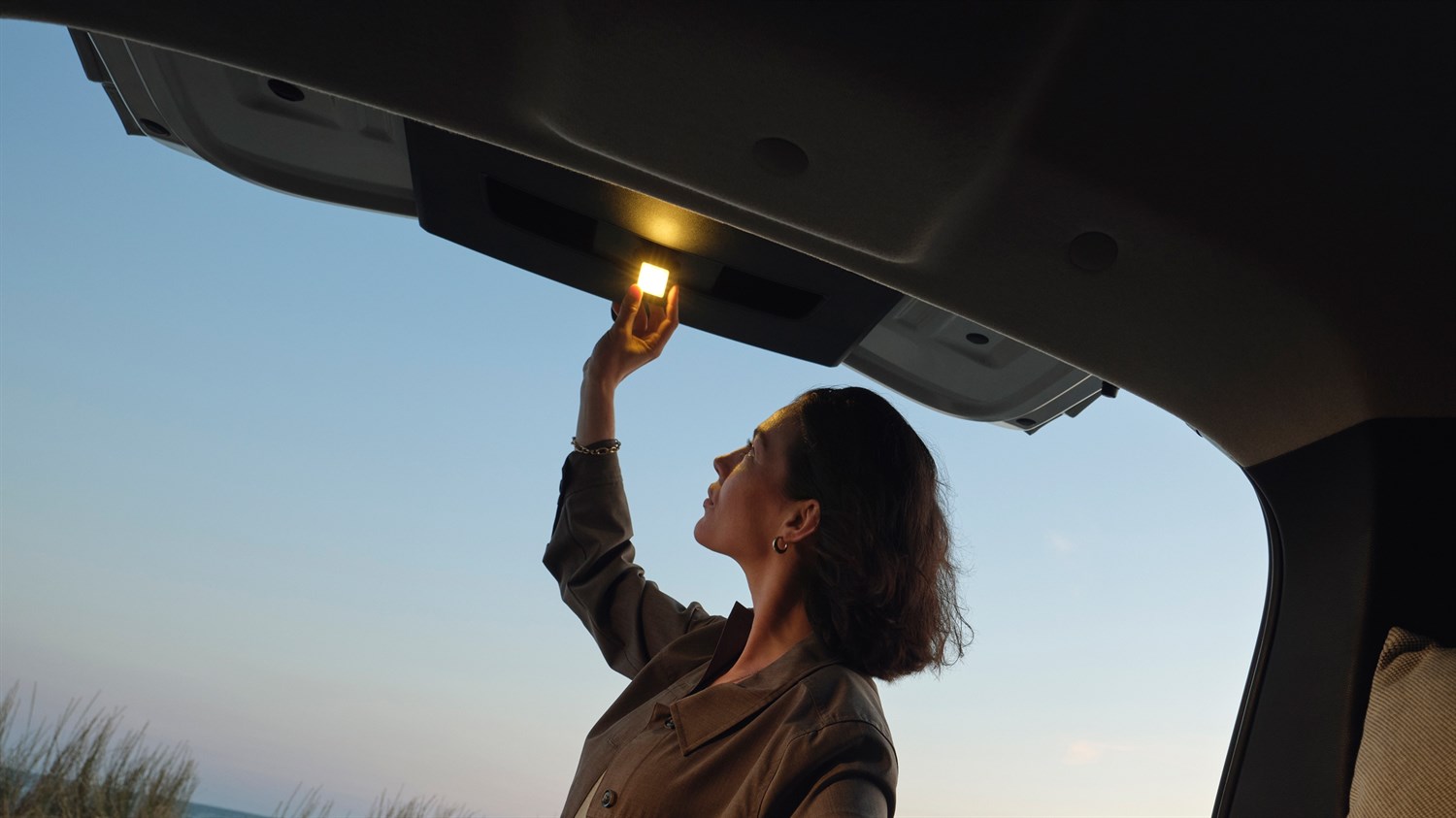Žena u vozilu Dacia Jogger pali unutarnje svjetlo na otvorenom prtljažnom prostoru pri zalasku sunca.
