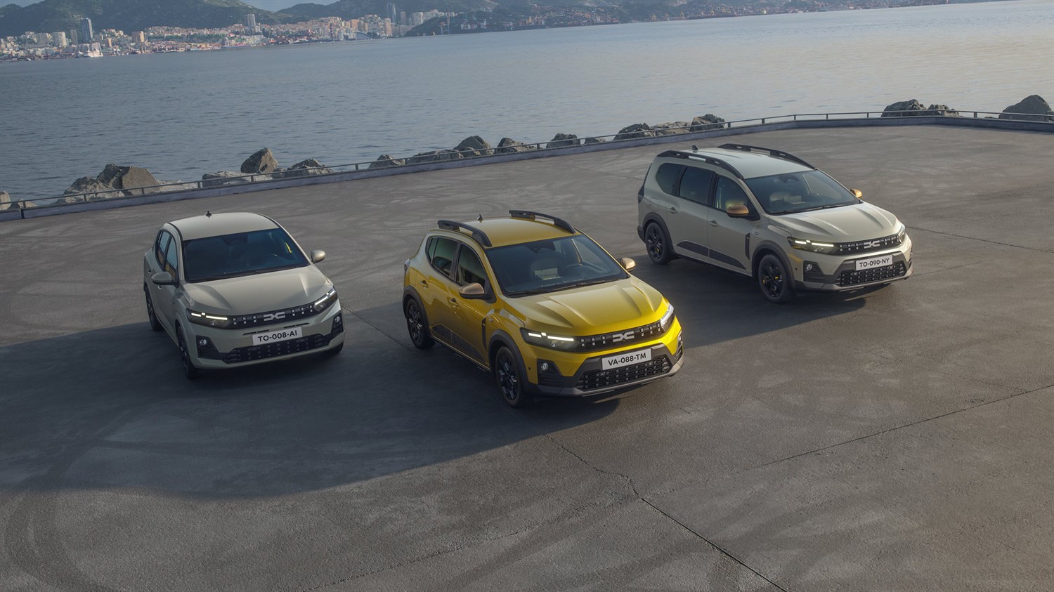 Tri vozila – Dacia Sandero, Dacia Sandero Stepway i vozilo Dacia Jogger – parkirana uz obalu s pogledom na more i grad u pozadini.