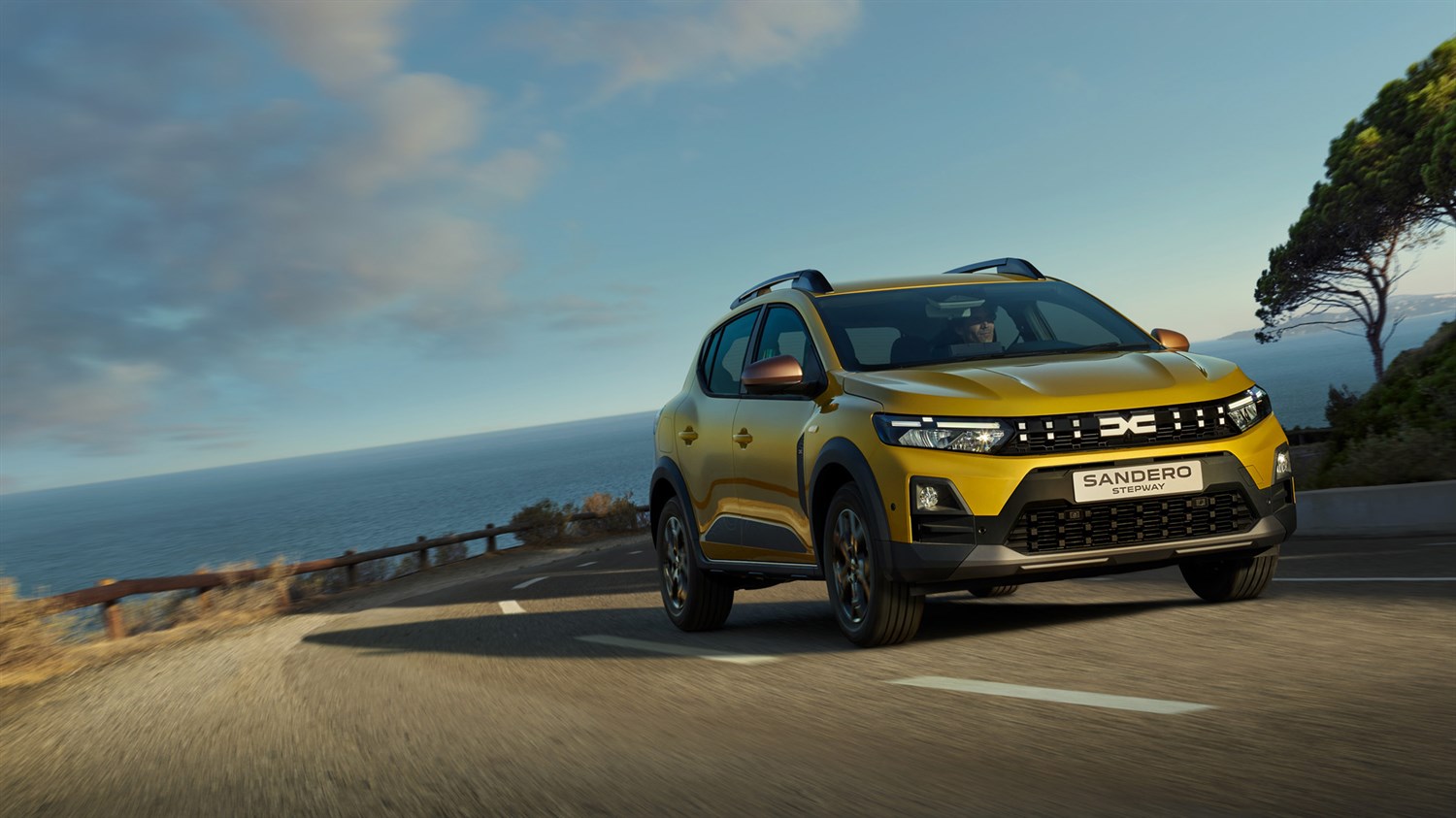 Vozilo Dacia Sandero Stepway u zlatnoj boji vozi se uz obalu s pogledom na more i plavo nebo u pozadini.