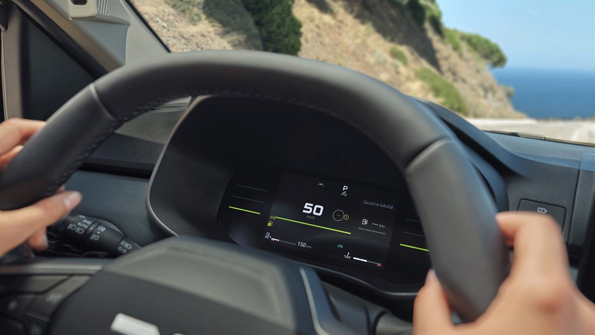 Pogled kroz upravljač vozila Dacia Jogger na digitalnu instrument ploču koja prikazuje brzinu vožnje, dok se u pozadini vidi obalna cesta i more.