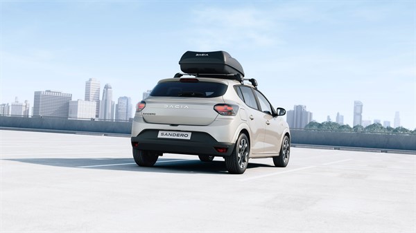 Vozilo Dacia Sandero s crnom krovnom kutijom na krovu vozi po otvorenom prostoru s gradom u pozadini.