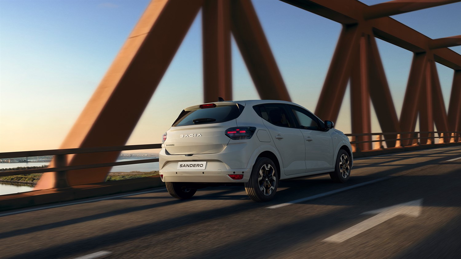 Vozilo Dacia Sandero u vožnji preko modernog mosta pri zalasku sunca, prikazano straga u pokretu.