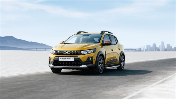 Žuti Dacia Sandero Stepway, pogled sprijeda, vozi cestom uz obalu, grad u pozadini.