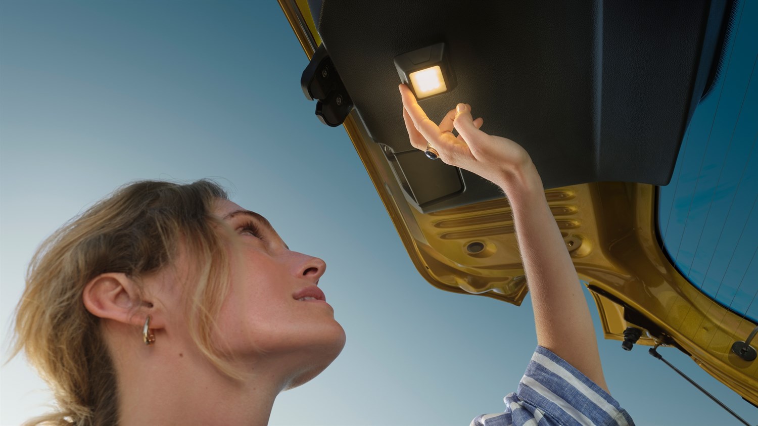 Žena podiže ruku prema unutarnjem svjetlu otvorenog prtljažnika vozila Dacia Sandero Stepway, snimljeno iz donjeg kuta prema nebu.