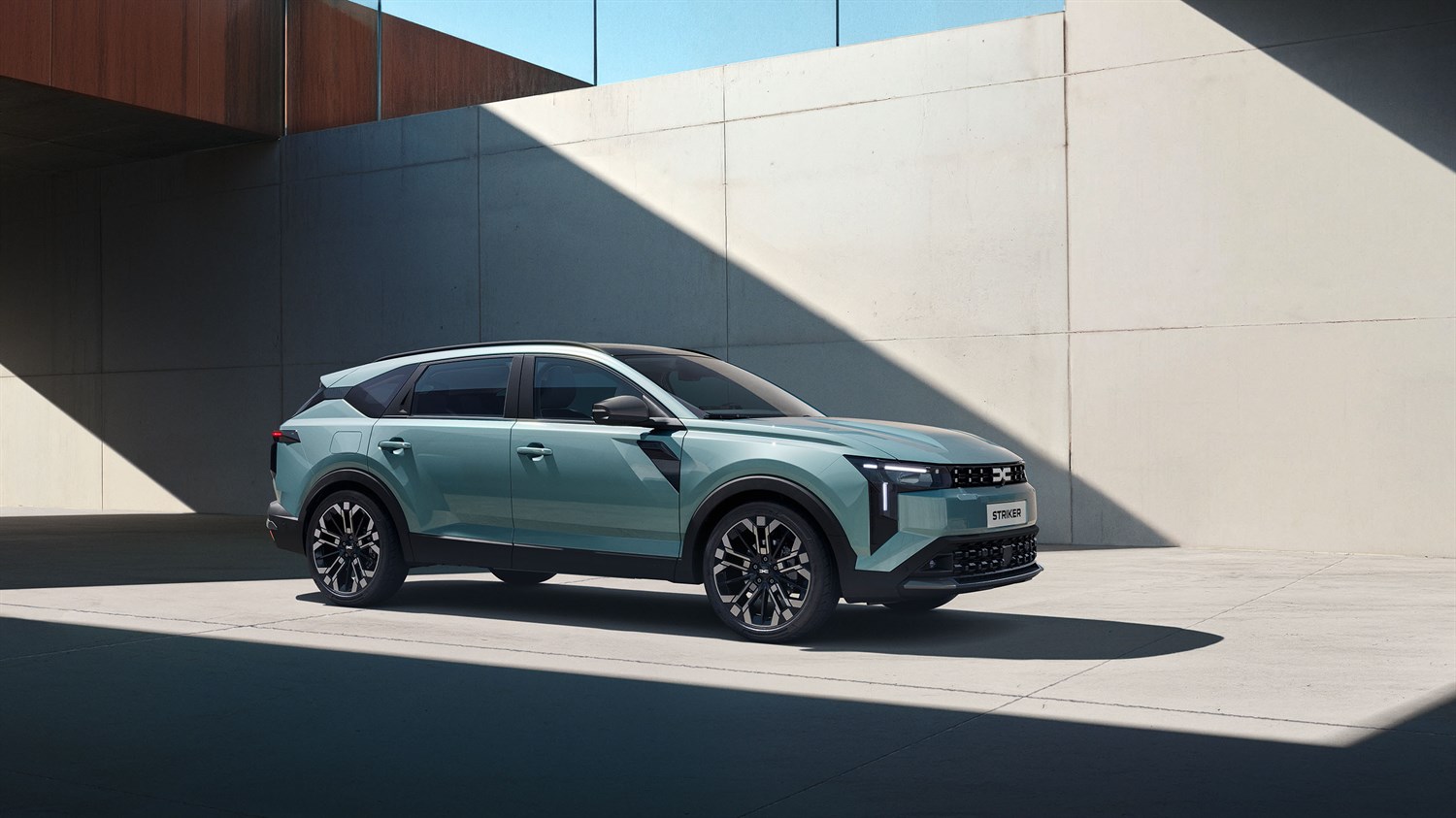 Vozilo Dacia Striker parkirano uz modernu betonsku zgradu, prikazano iz bočnog kuta s naglašenim linijama karoserije i velikim aluminijskim naplatcima, dok preko zida pada snažna dijagonalna sjena koja ističe dizajn vozila