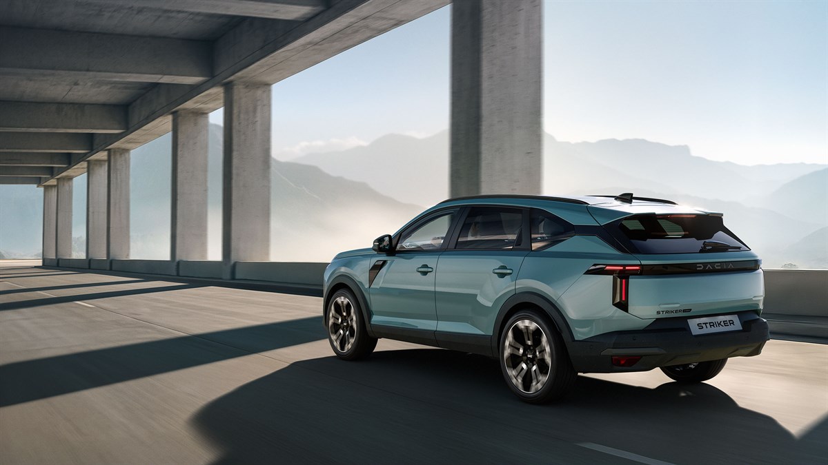 vozilo Dacia Striker prikazano iz stražnje bočne perspektive dok vozi kroz moderan betonski prolaz s velikim stupovima, uz planinski krajolik u pozadini i naglašena stražnja LED svjetla te dinamične linije vozila