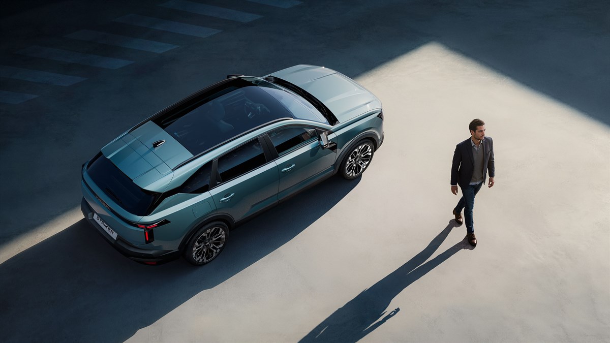 vozilo Dacia Striker prikazano iz gornje perspektive parkirano na otvorenom prostoru, s panoramskim krovom i naglašenim linijama karoserije, dok osoba prolazi pored vozila i stvara dugu sjenu na tlu