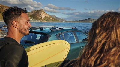 Dvoje ljudi u neoprenima stoje kraj mora, jedan drži surf dasku, a iza njih je parkiran zeleni automobil uz obalu, s planinama u pozadini.