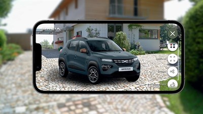 Na slici je prikazan ekran pametnog telefona na kojem se vidi aplikacija proširene stvarnosti (AR). Na ekranu se prikazuje automobil marke Dacia Spring, koji izgleda kao da je parkiran ispred kuće na popločanom prilazu.